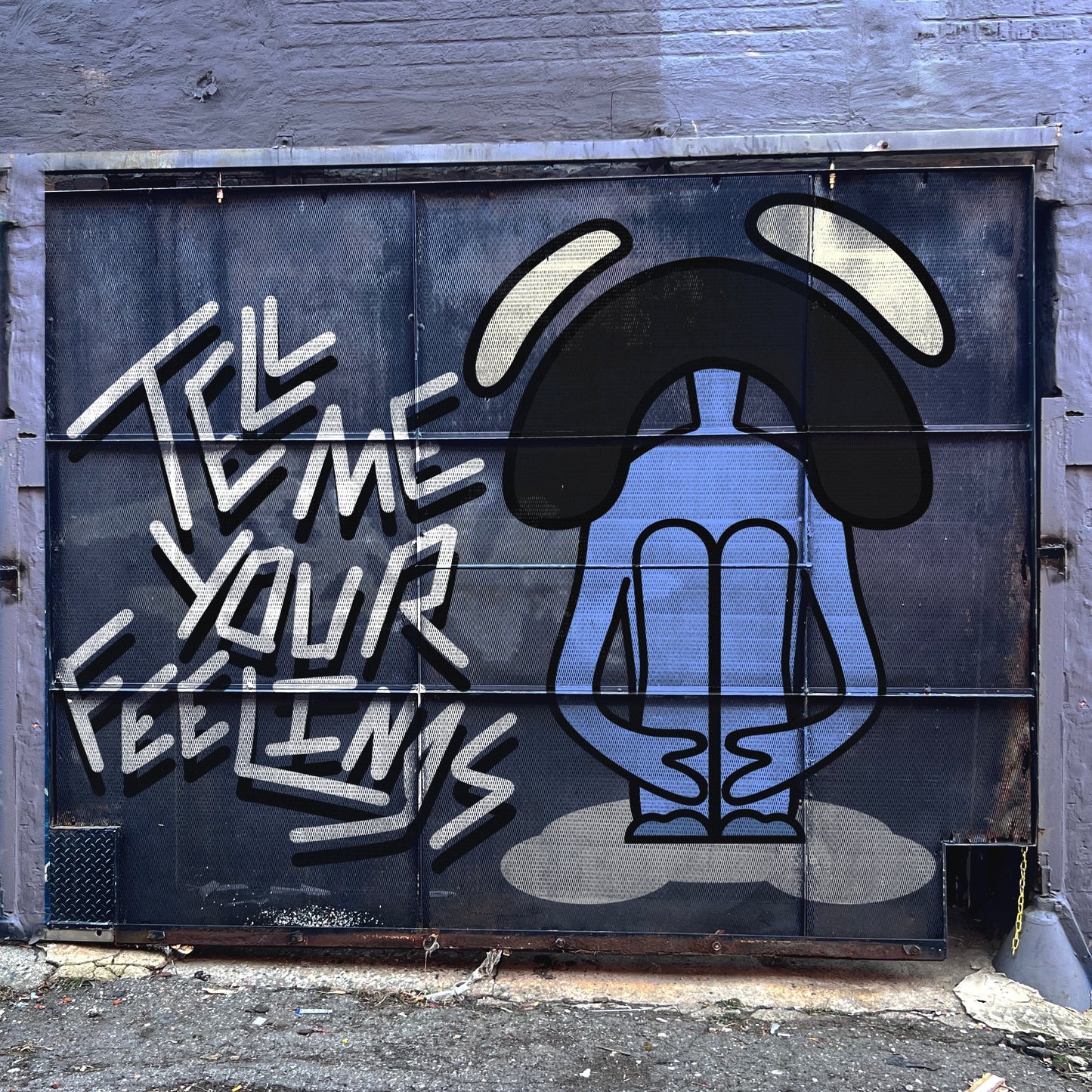 Street art mural on a metal door showing a stylized blue figure sitting cross-legged with elongated arms resting on their knees and a bowed head, surrounded by large black curved shapes resembling ears or antennae. Bold graffiti text on the left reads “TELL ME YOUR FEELINGS” in white, angular letters. The setting appears to be an urban alley with a concrete ground and weathered walls.