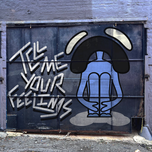 Street art mural on a metal door showing a stylized blue figure sitting cross-legged with elongated arms resting on their knees and a bowed head, surrounded by large black curved shapes resembling ears or antennae. Bold graffiti text on the left reads “TELL ME YOUR FEELINGS” in white, angular letters. The setting appears to be an urban alley with a concrete ground and weathered walls.